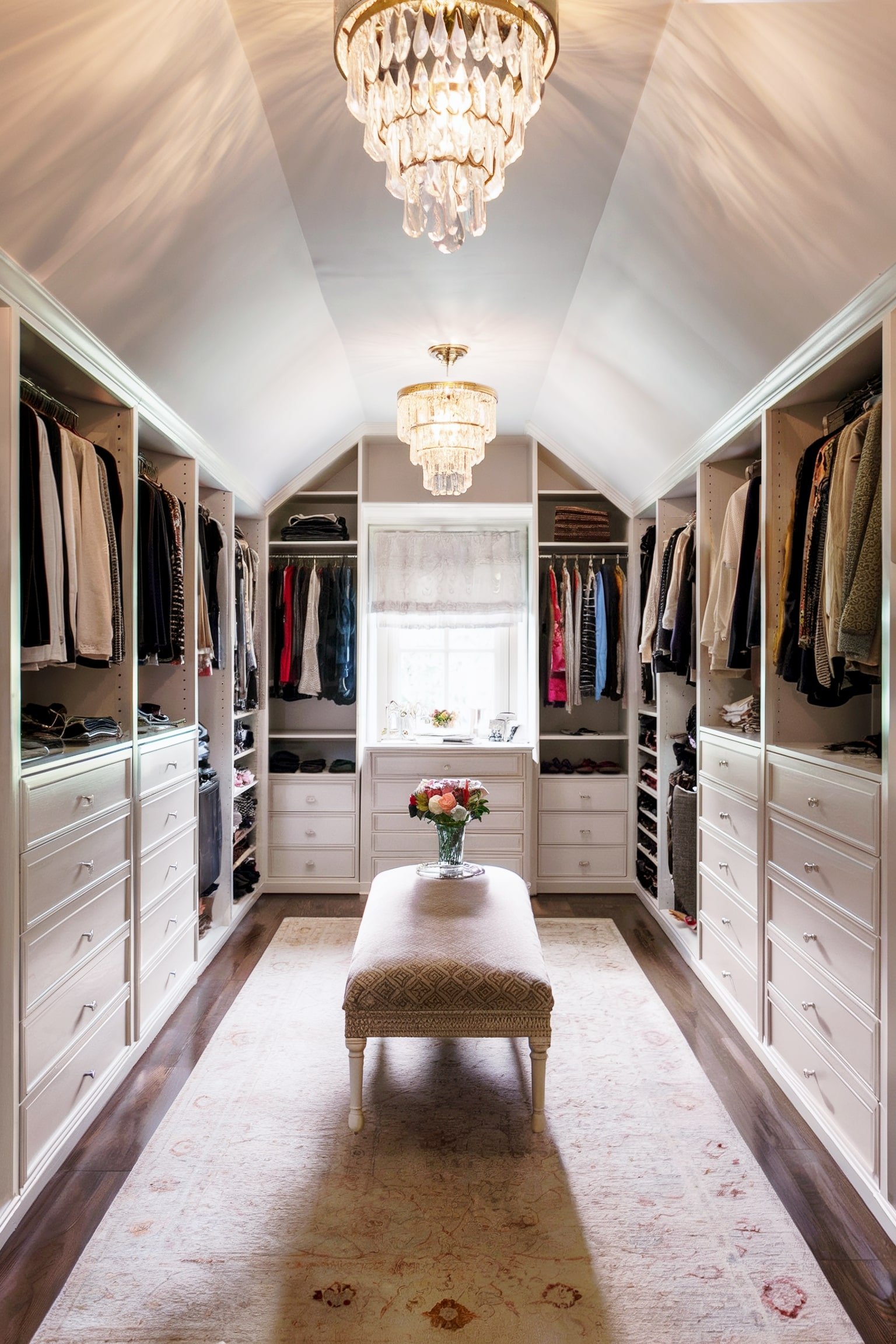 White closet with hanging clothes, chandeliers, and center bench