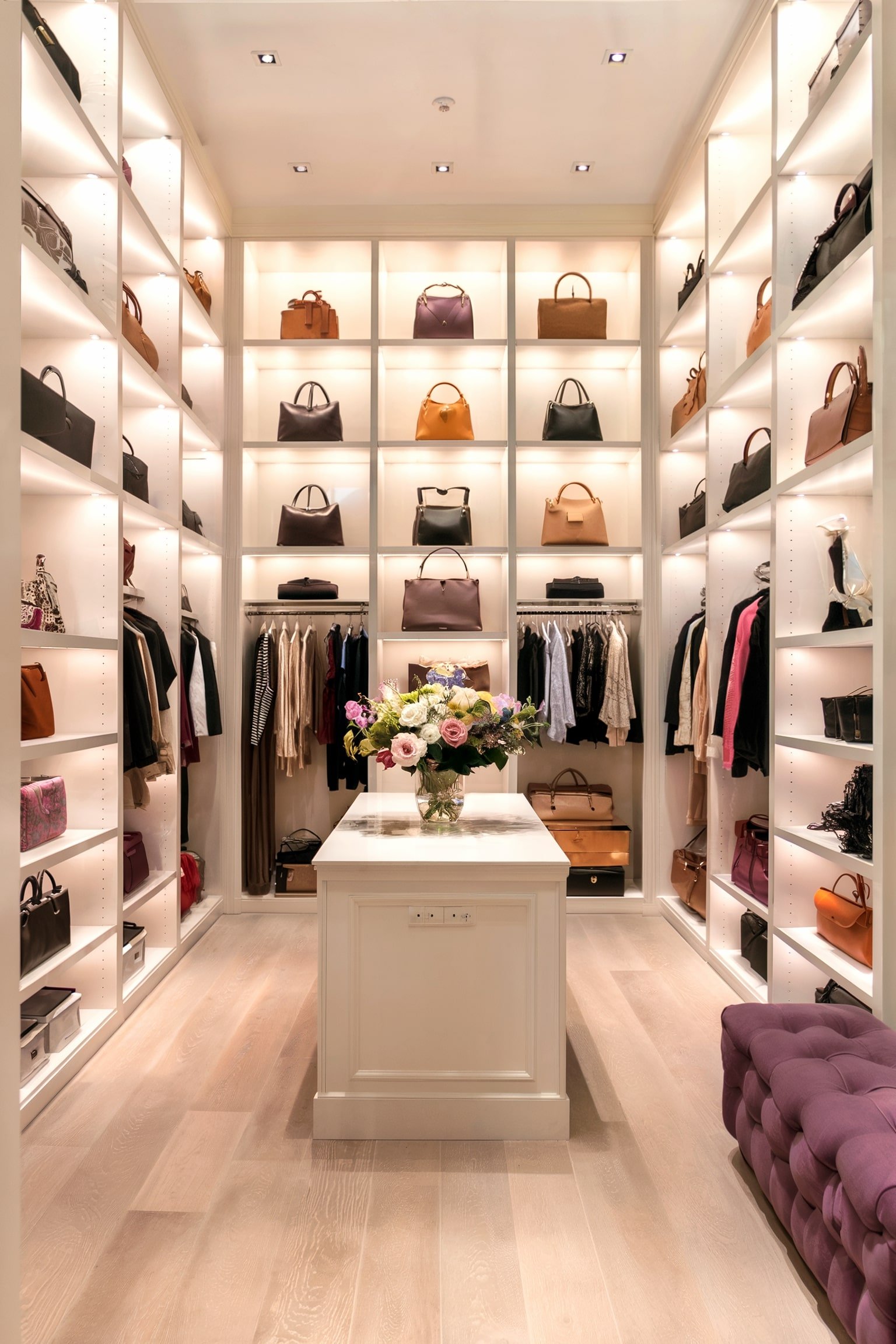 Well-organized closet featuring high, illuminated shelves with handbags, a central island, and decorative flowers.