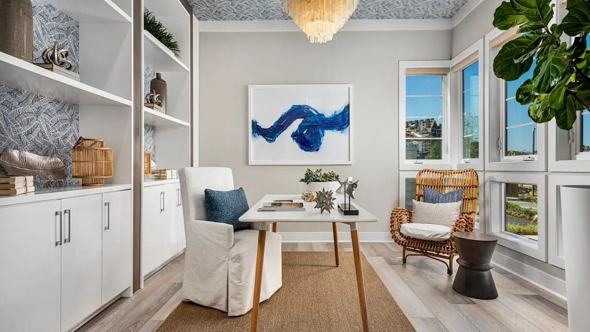 A home office with windows, a shelf, cabinets, a chandelier, a painting, chairs, and a desk.