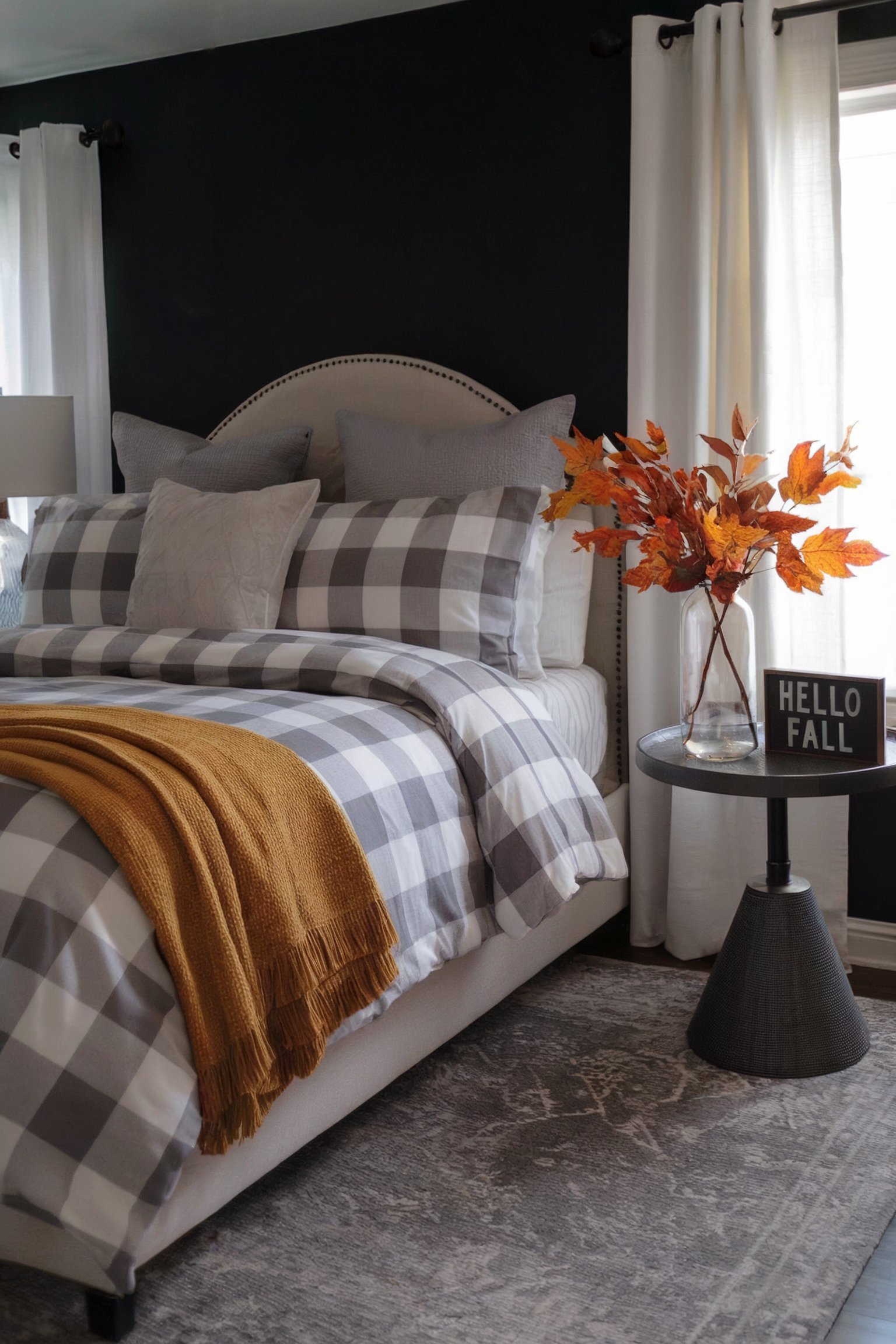 A bedroom featuring a black accent wall, gray buffalo check bedding, and fall leaves in a vase with a "Hello Fall" sign on the bedside table