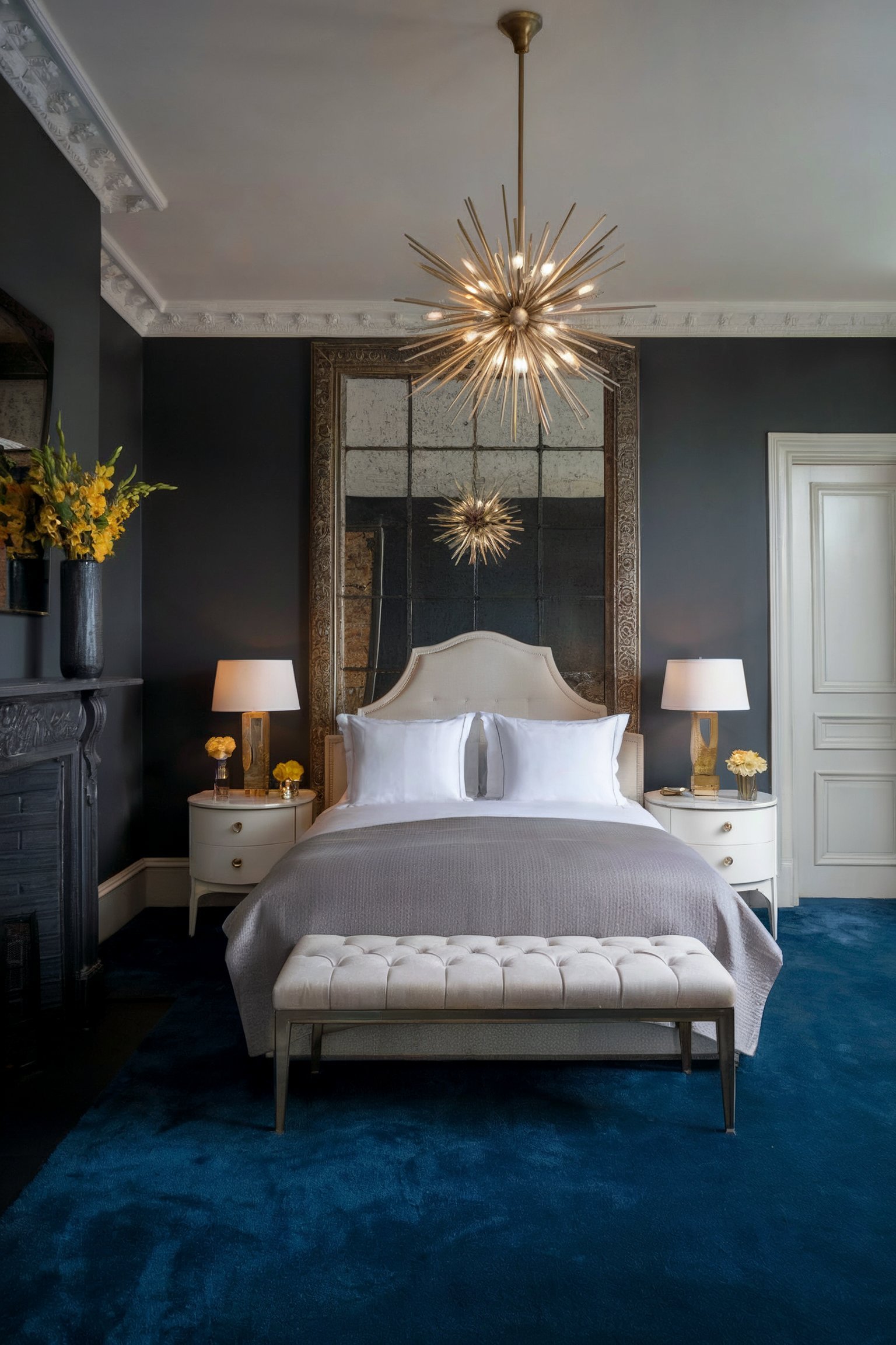 A luxurious bedroom with dark walls, a tufted beige headboard, blue carpet, and a large, ornate chandelier overhead
