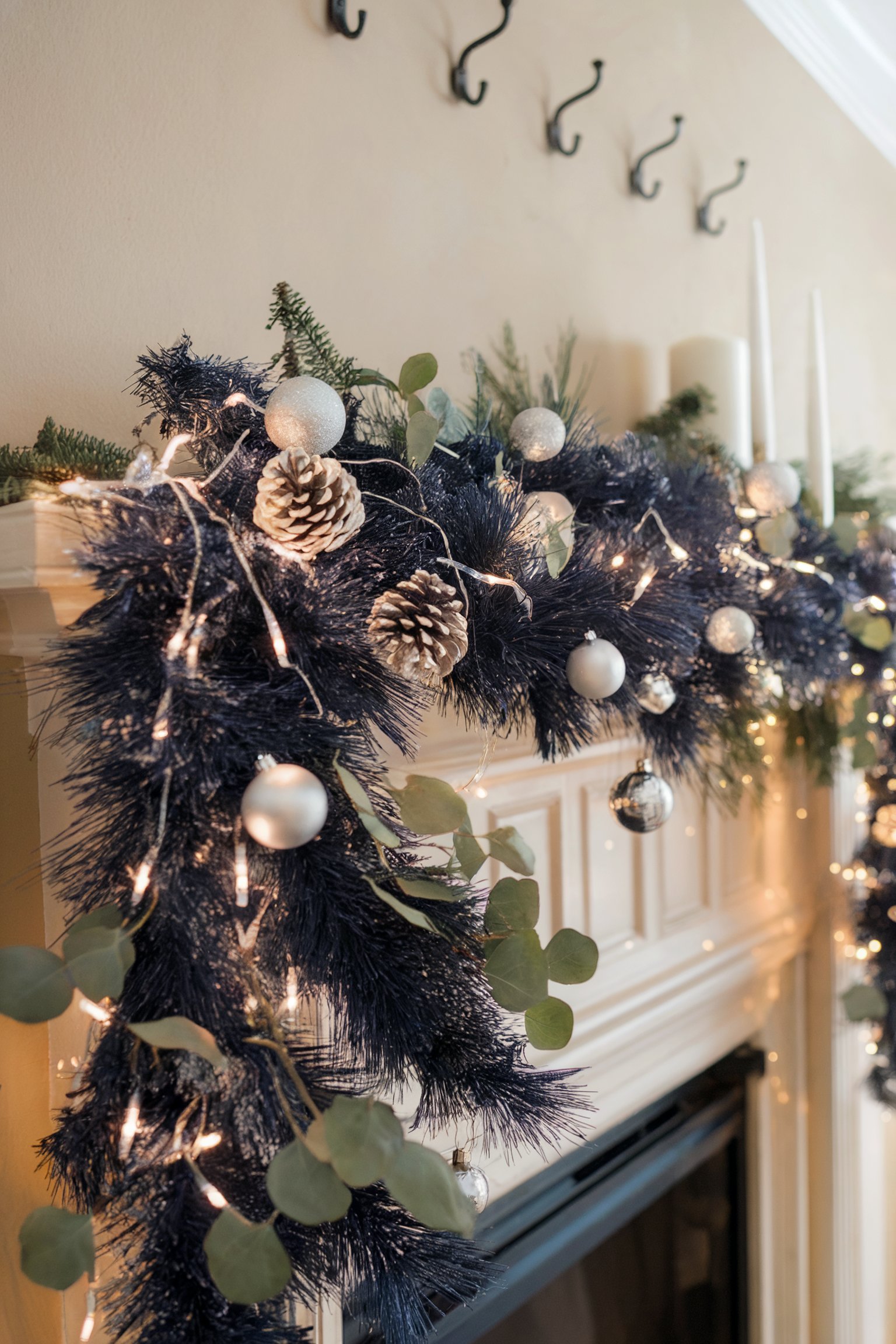 A festive fireplace mantel decorated with dark blue garland, silver ornaments, pinecones, eucalyptus leaves, and white string lights