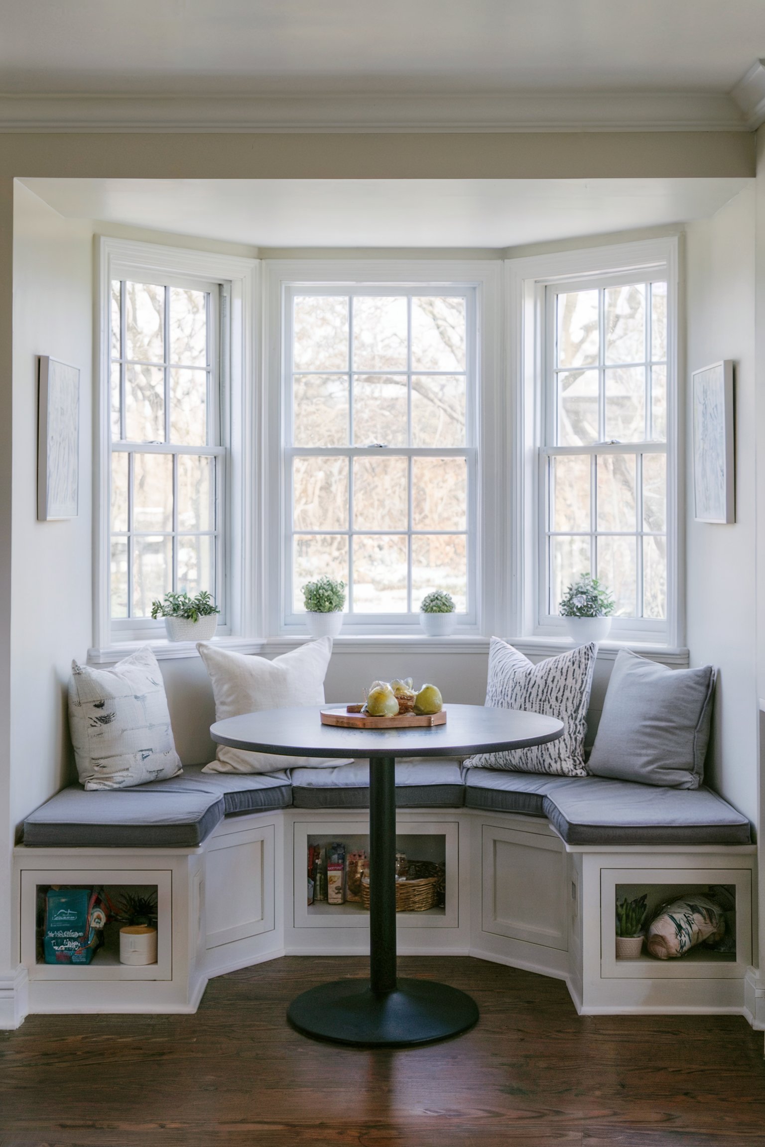 A bright kitchen nook with a round table, surrounded by bay windows and cozy seating with built-in storage beneath