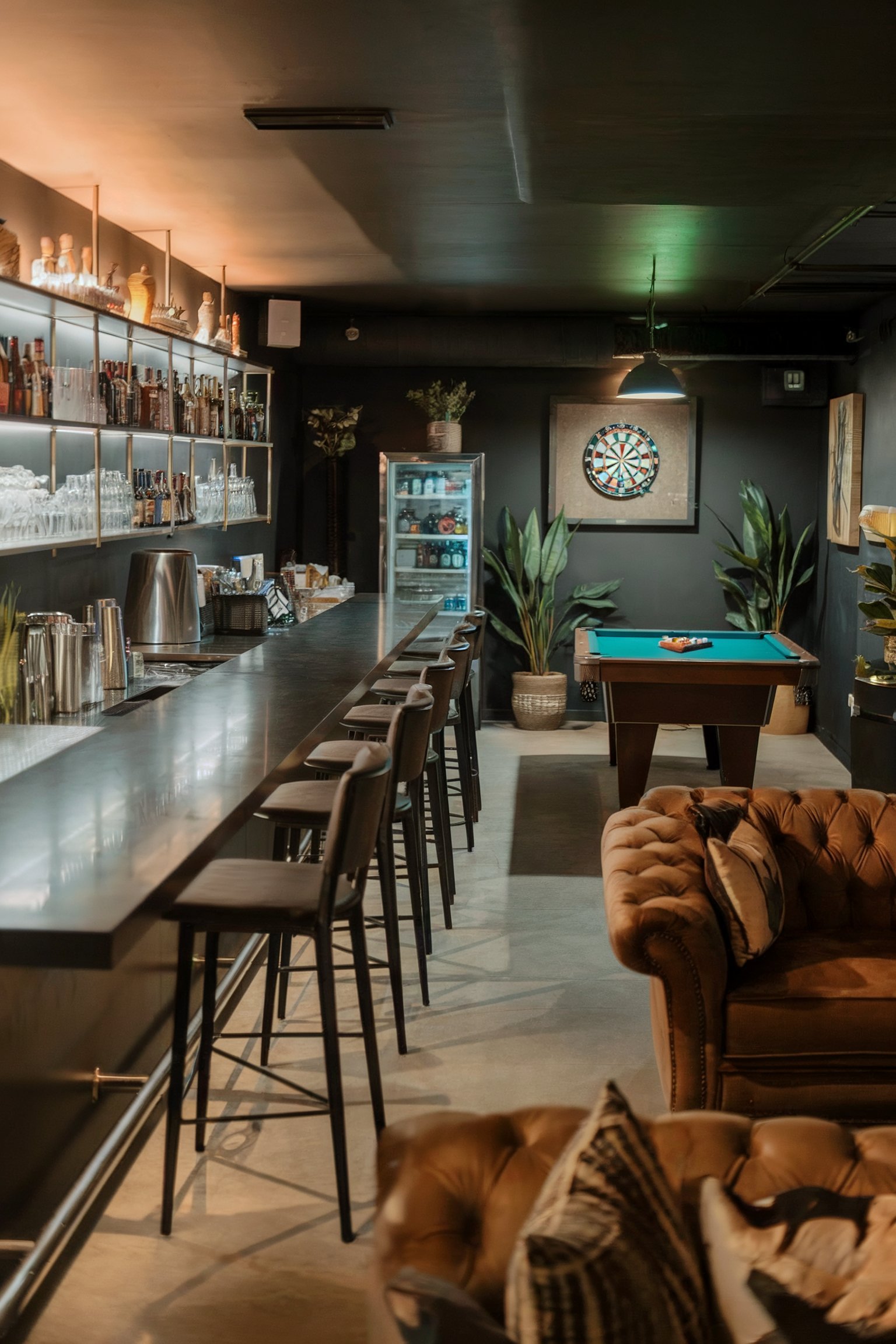 A modern basement bar featuring a long counter with barstools, a pool table, a dartboard, and industrial lighting
