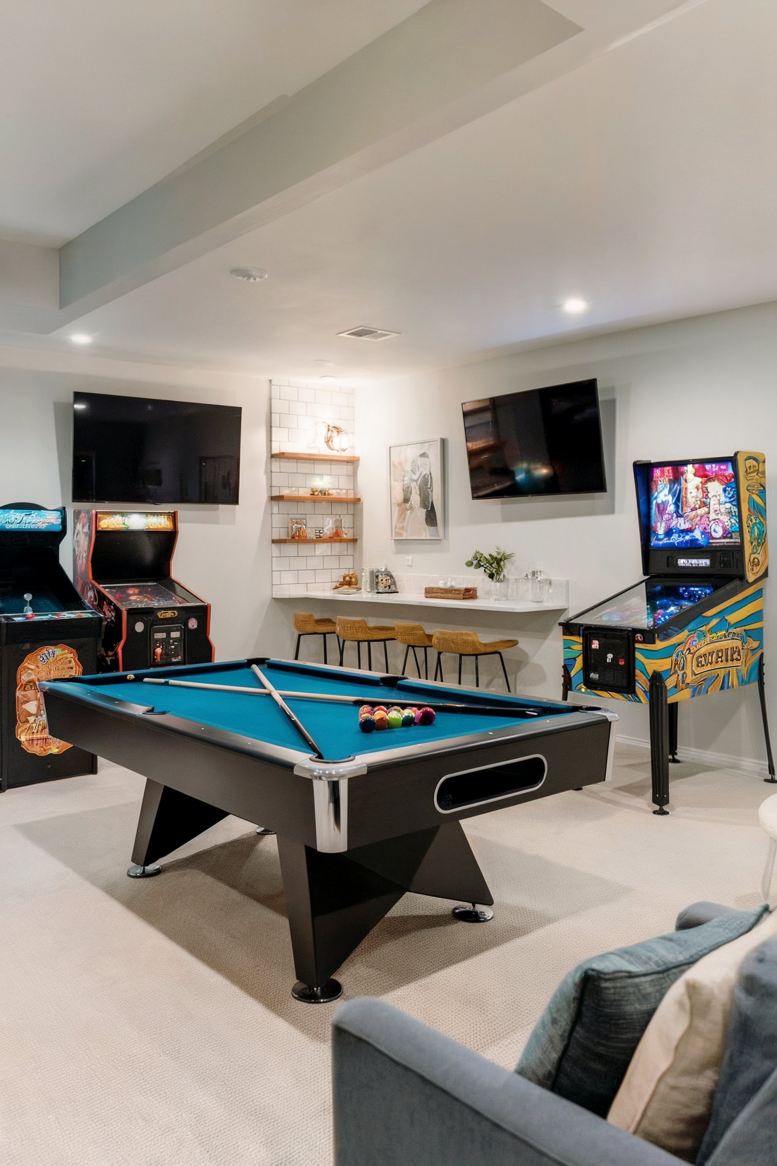 A sleek basement game room with a pool table, arcade games, and pinball machine, with two wall-mounted TVs and a small bar area