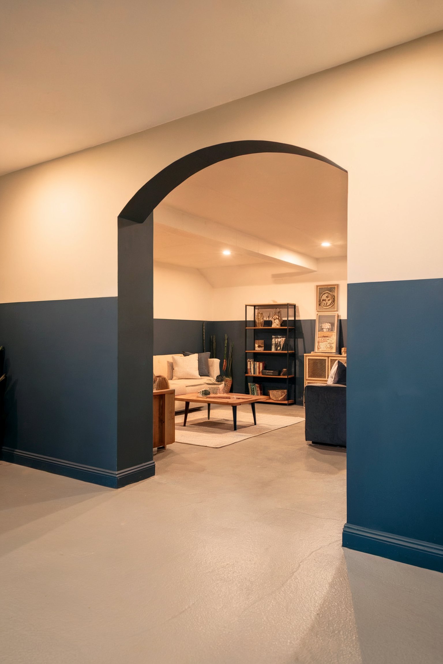A modern basement room with dark blue and white walls, an arched entrance, and a seating area with a beige couch and a bookshelf