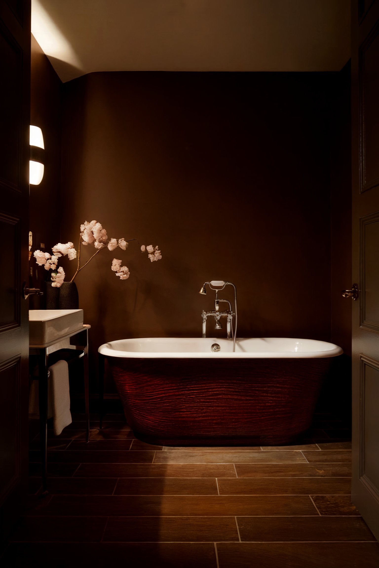 A dimly lit bathroom with a deep brown wall, featuring a white freestanding bathtub with a rich wooden exterior, and a modern faucet, alongside a simple vanity