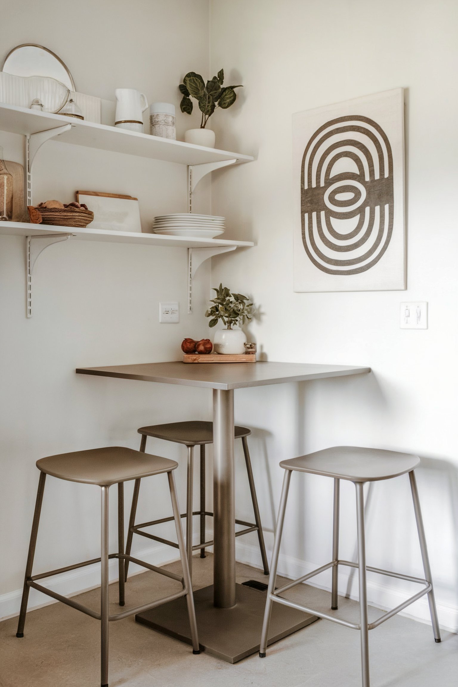 A minimalist kitchen nook with a small high-top table, three stools, and neutral shelving decor
