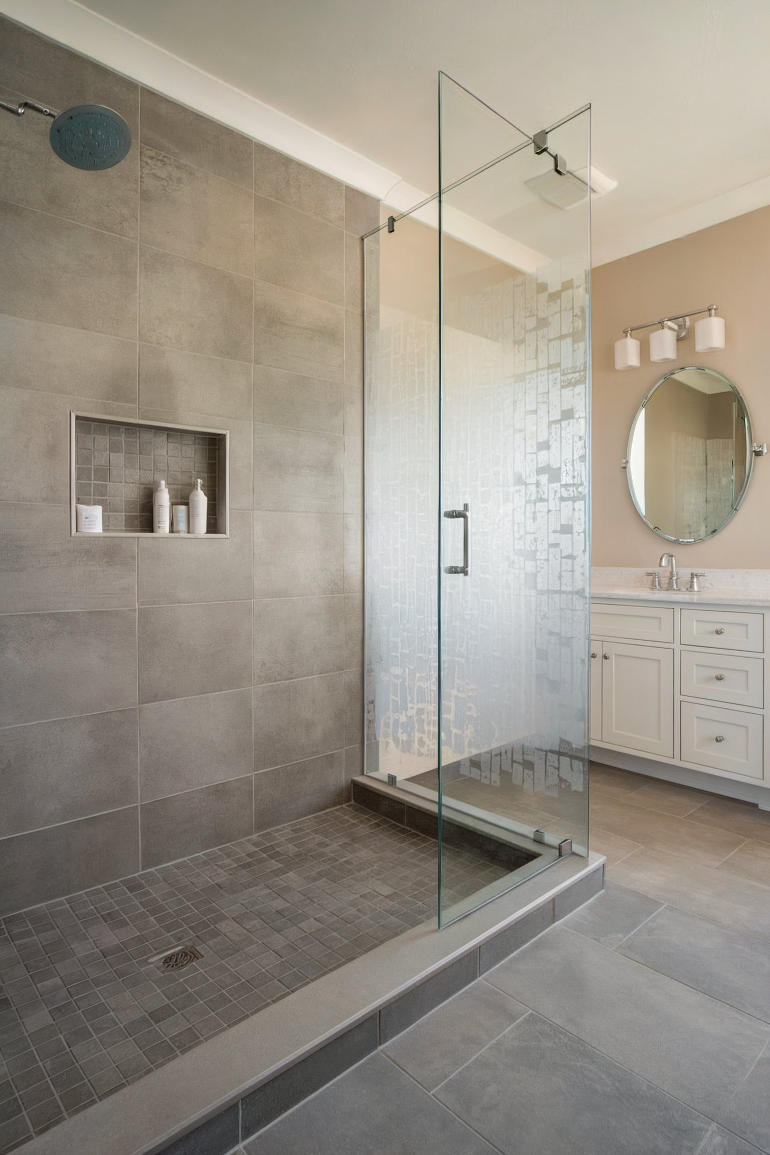 Gray-tiled shower with built-in shelf, glass door, and white vanity with a round mirror