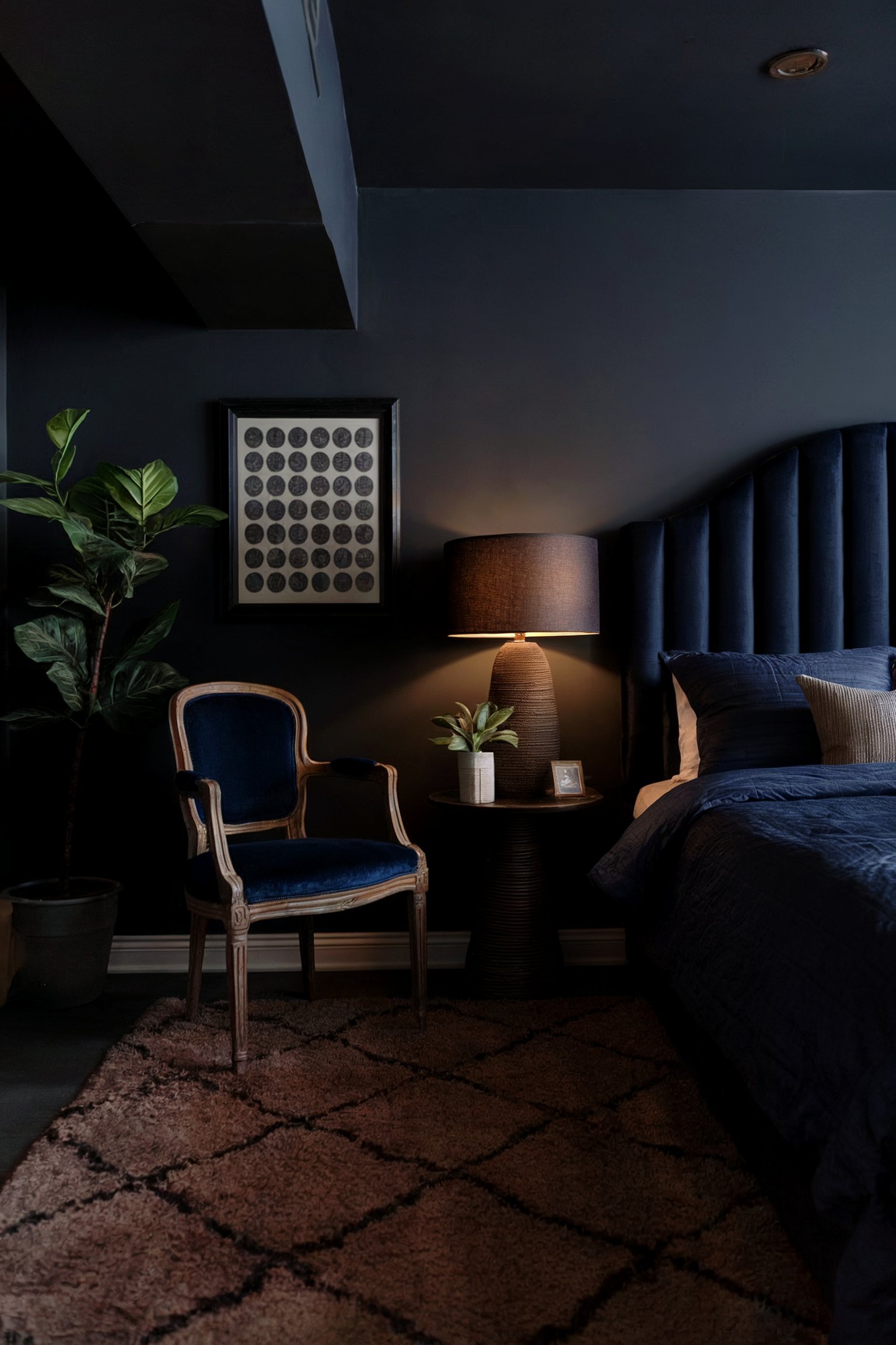 A moody bedroom with dark walls, a navy headboard, and a warm lamp, accompanied by a vintage-style armchair and a large potted plant