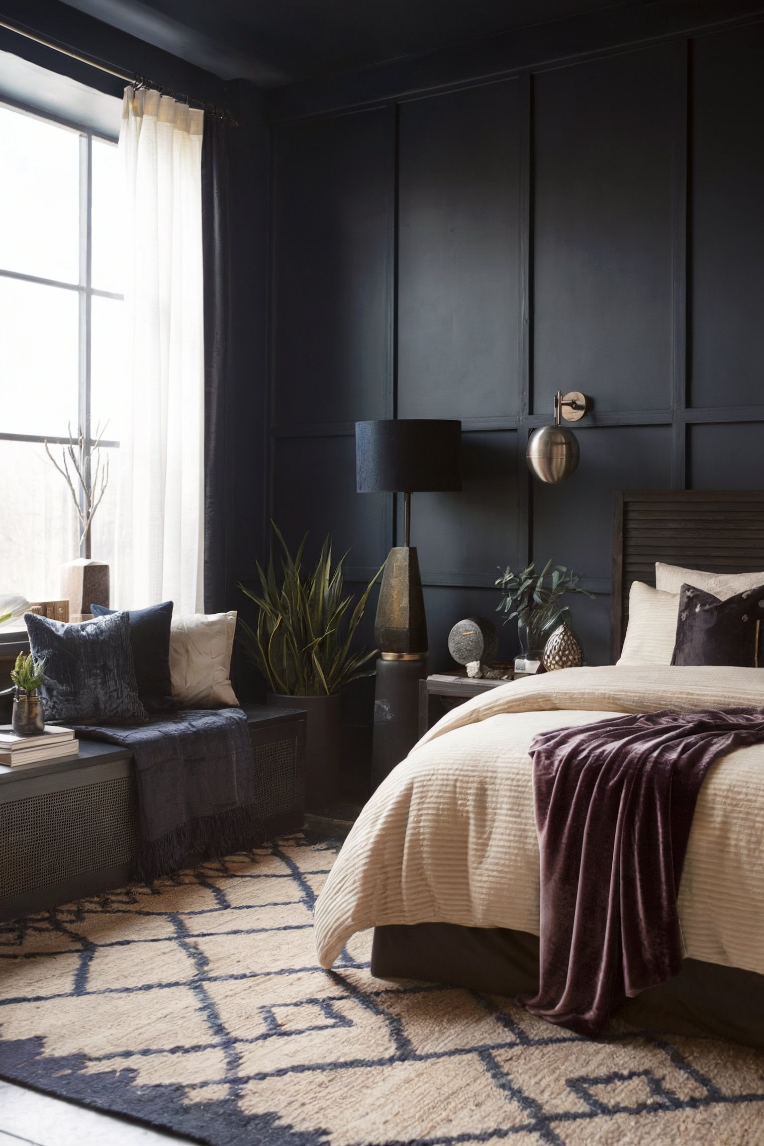 A modern, moody bedroom with dark blue walls, cream bedding, and contrasting geometric-patterned rug, accented by a floor lamp and indoor plants