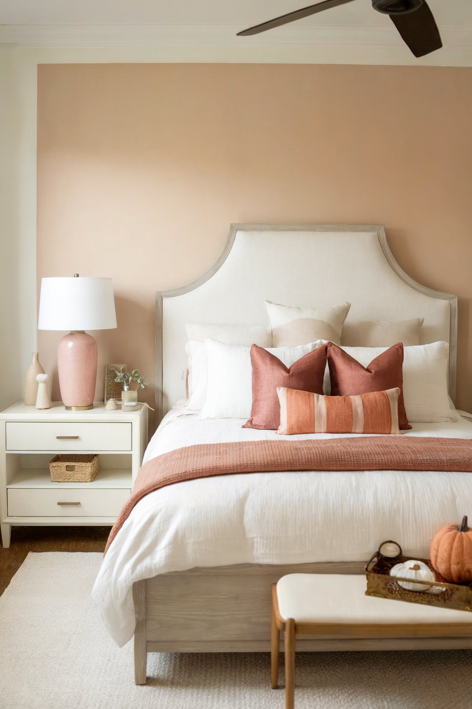 A soft, light-filled bedroom with a beige upholstered bed, peach accent pillows, and a wooden bench