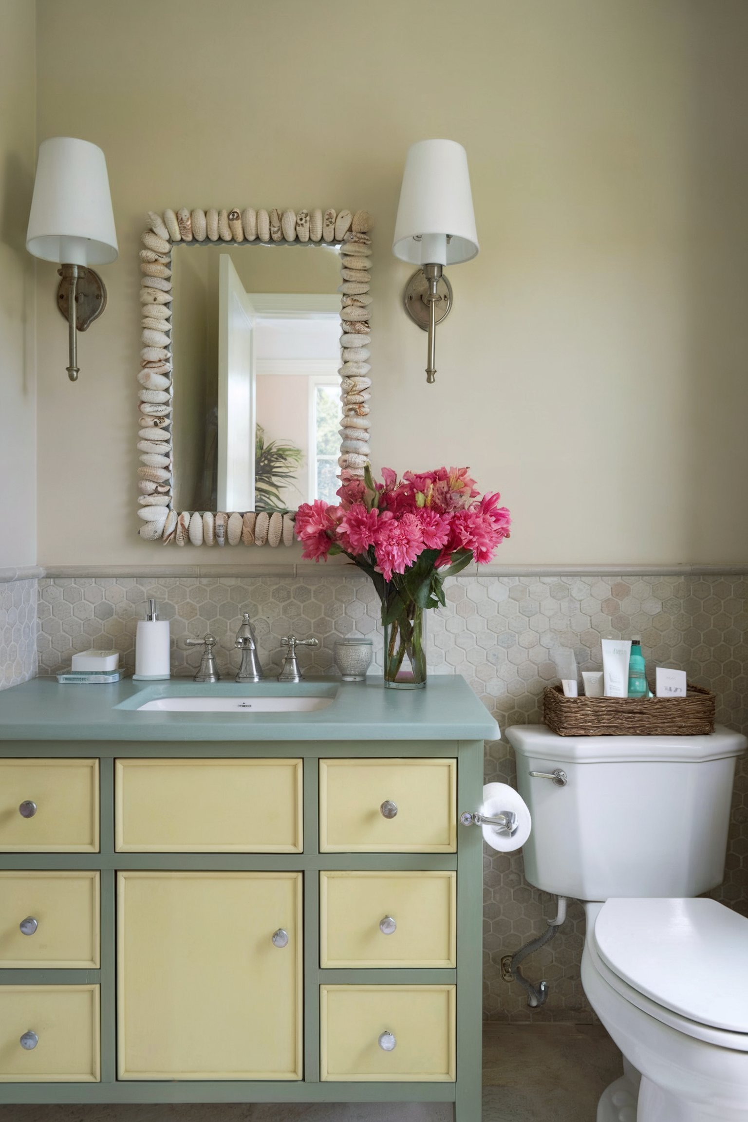 A pastel yellow and light green vanity is paired with a seashell mirror and pink flowers, creating a soft, coastal-inspired bathroom atmosphere