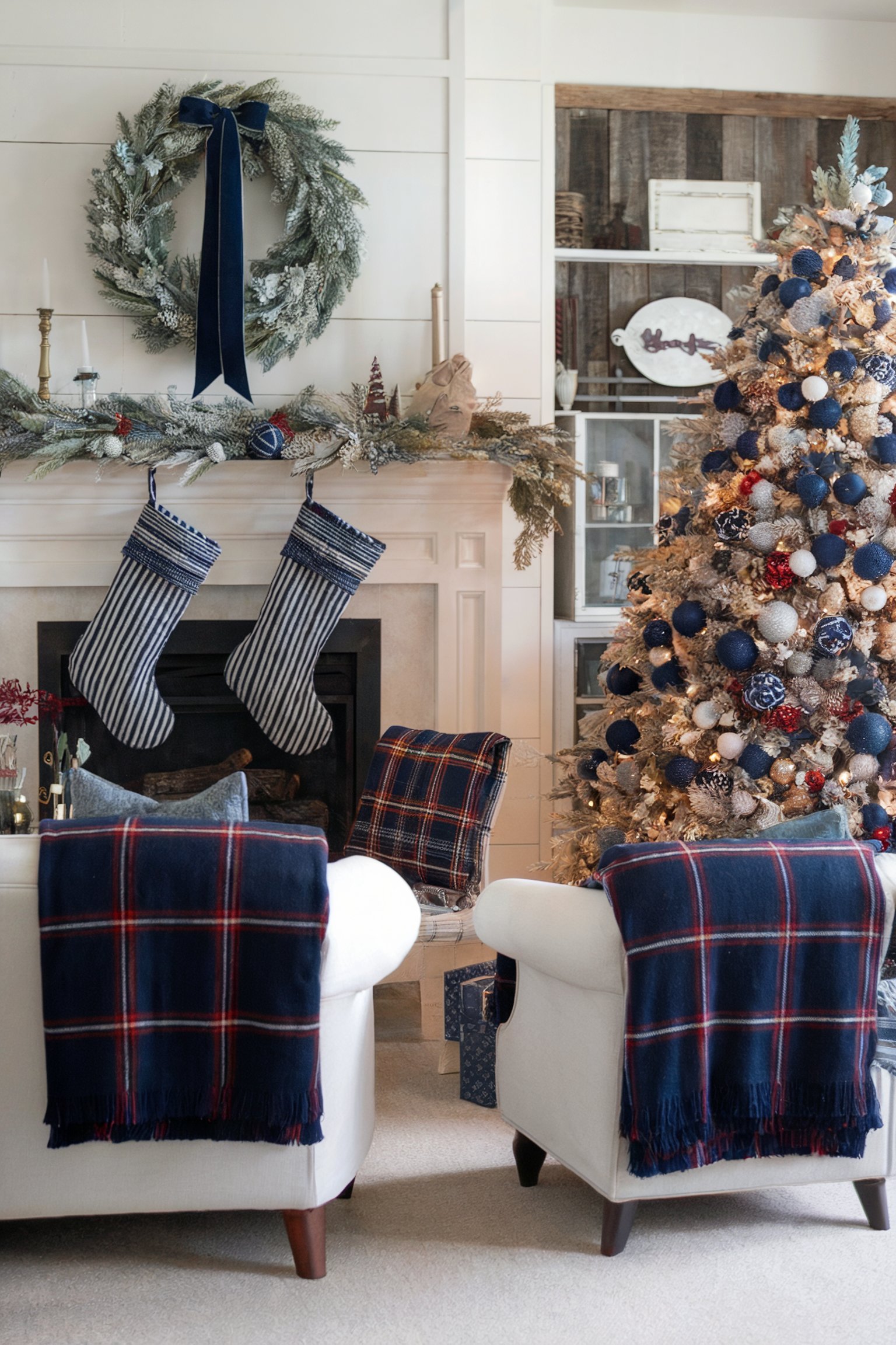 A cozy farmhouse-style Christmas setup with a decorated mantel and tree. The mantel features a large frosted wreath with a navy blue ribbon, hanging stockings, and greenery