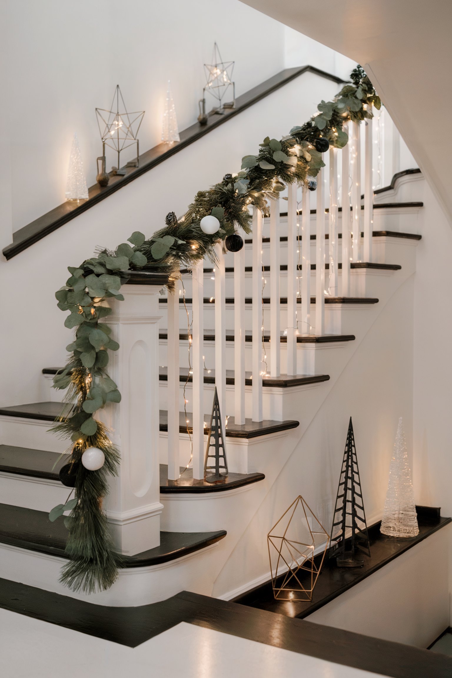 Modern staircase wrapped in eucalyptus garland and string lights, with decorative Christmas trees