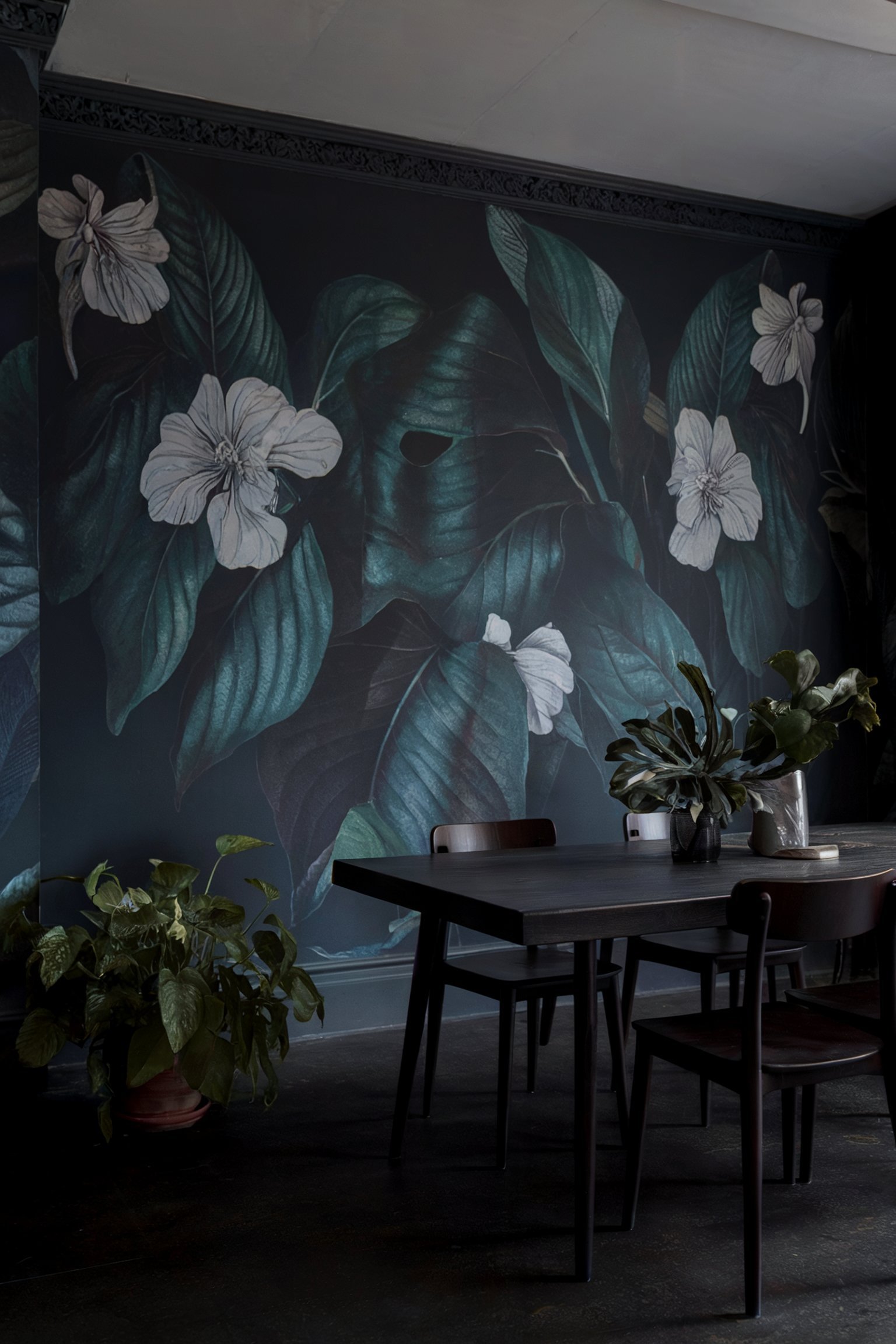Dark, moody dining room with dramatic large-scale foliage wallpaper, a black dining table, dark chairs, and potted greenery