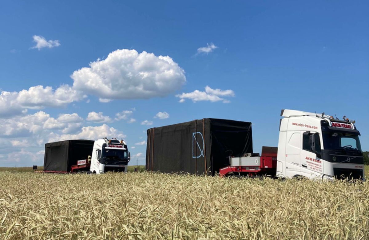 Two large semi-trailer trucks transporting what appear to be modular building sections across a field of tall crops, likely wheat or a similar grain