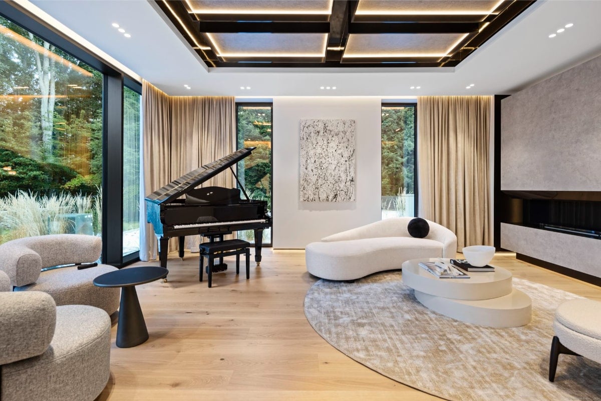 Living room with floor-to-ceiling windows offering views of greenery. A grand piano is prominently featured, and the furniture includes a curved white sofa and other light-colored seating. The ceiling has an interesting design with dark wood beams.