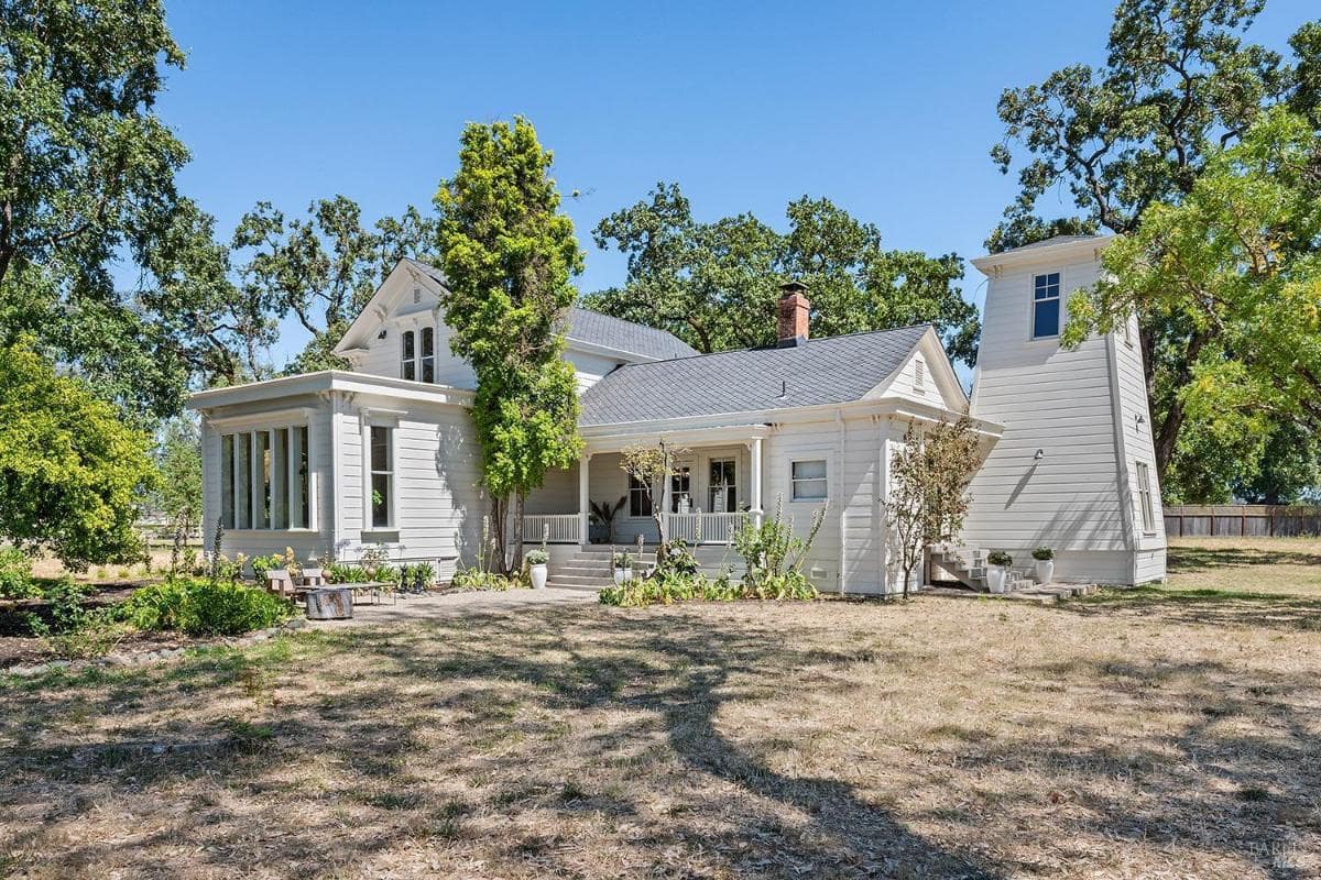 Exterior of white house with large windows, wraparound porch, and surrounding trees.