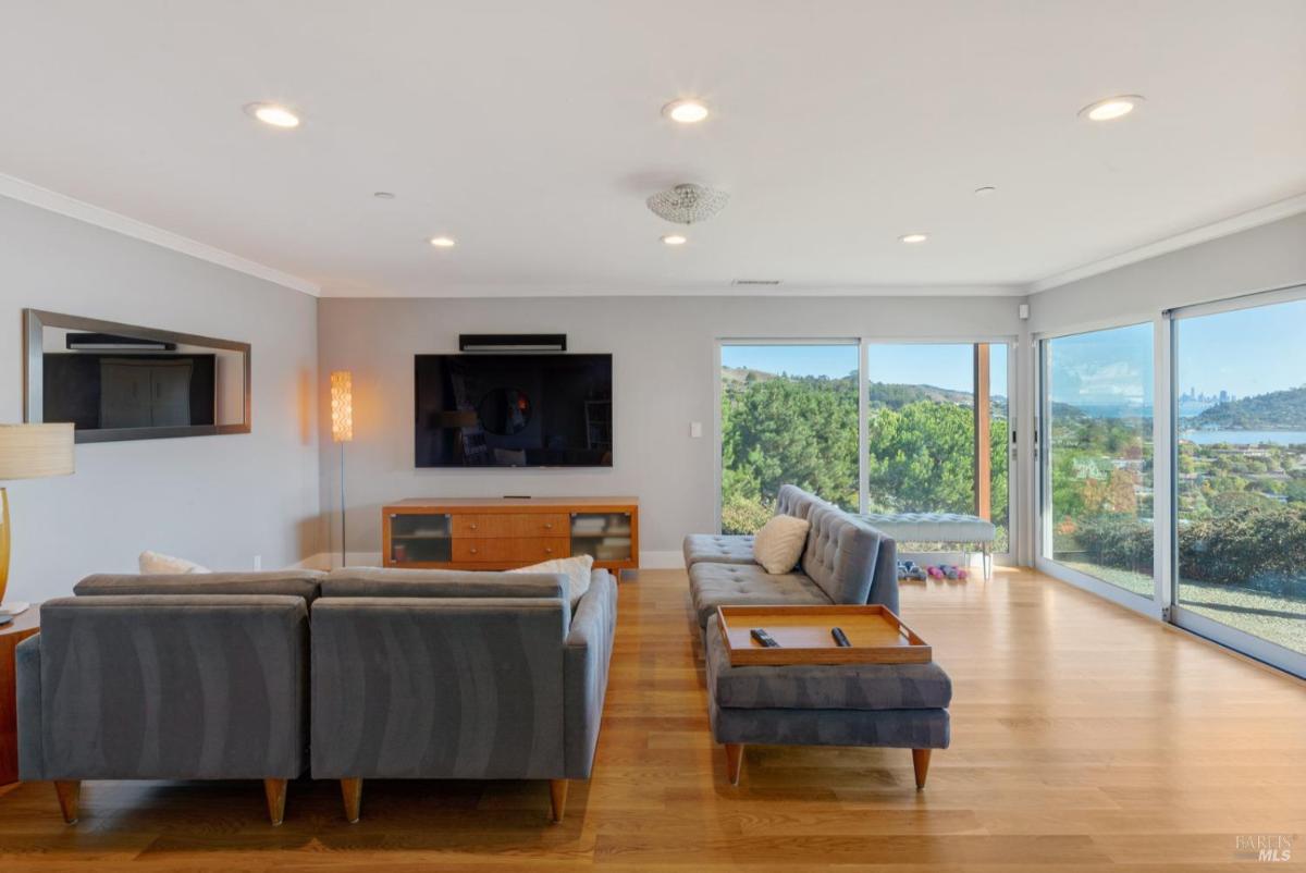 A family room with a gray sofa faces a large TV and has expansive windows offering a view of the outdoors.