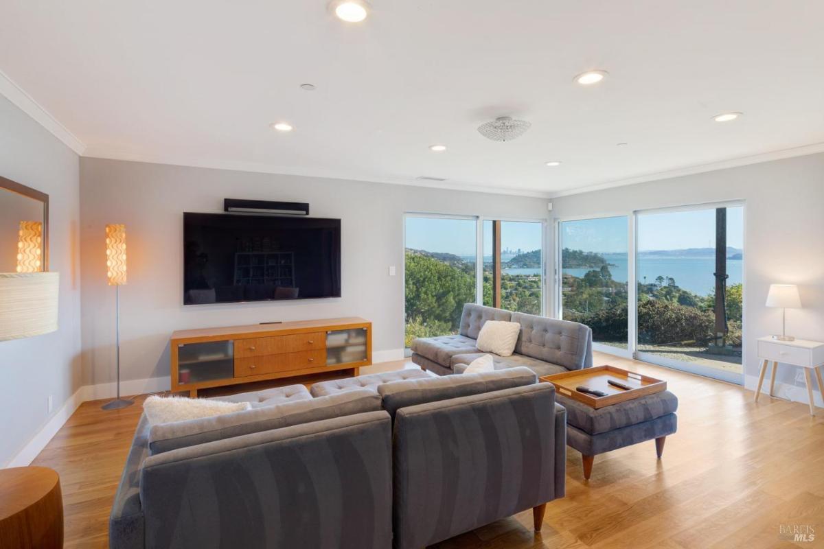 A family room with a gray sofa faces a large TV and has expansive windows offering a view of the outdoors.