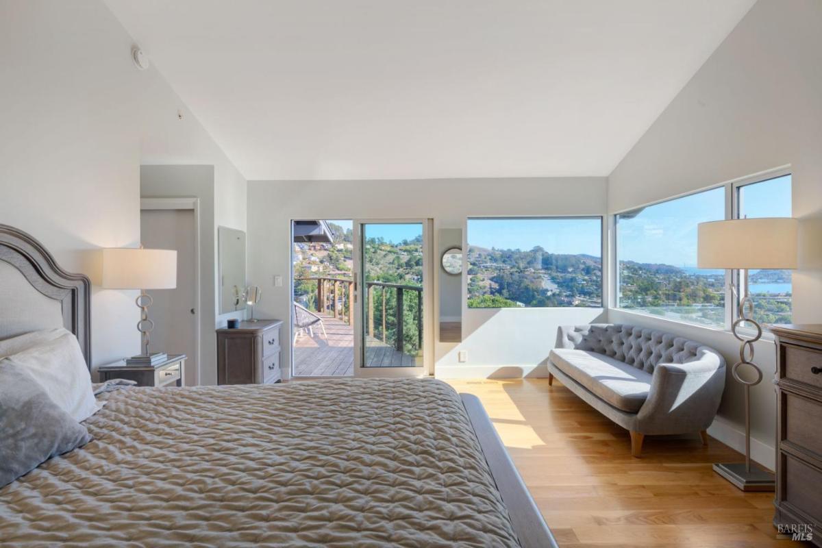 A bedroom features a large bed with a cozy quilt, a stylish gray sofa, and windows that provide a beautiful view of the landscape outside.