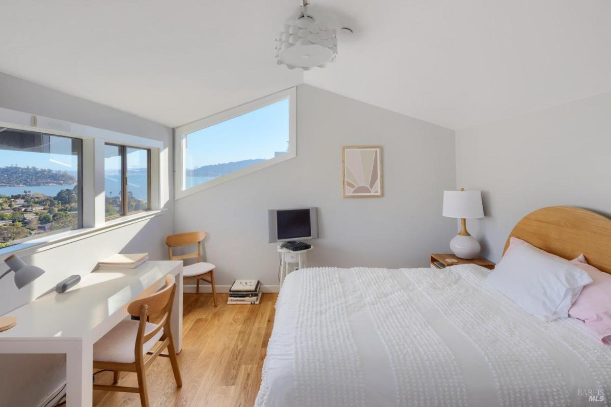 A bedroom with a bed, desk, two chairs, lamp, and a small TV. Large windows show a view of water and hills.