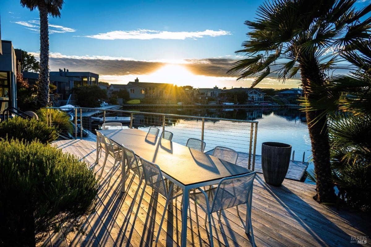 An outdoor dining area with a metal table and chairs beside a waterfront at sunset. 
