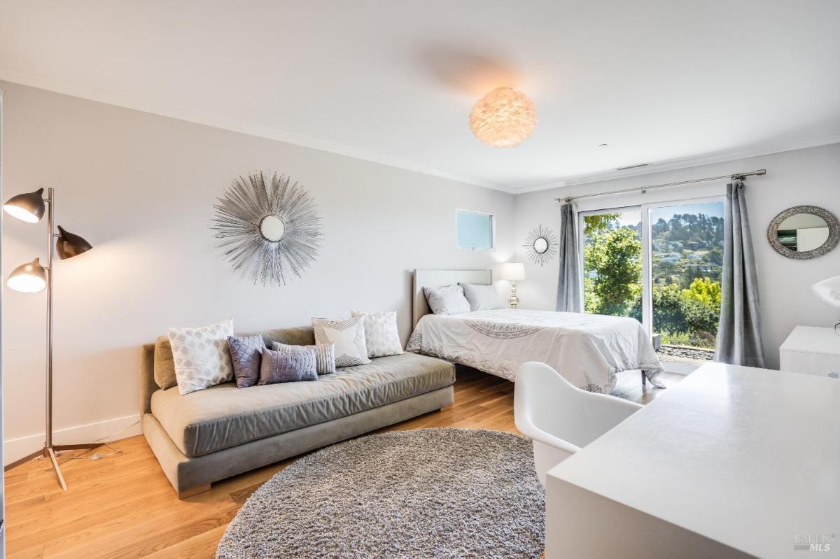 A bedroom with a bed, sofa, desk, floor lamp, and sliding glass doors overlooking greenery.