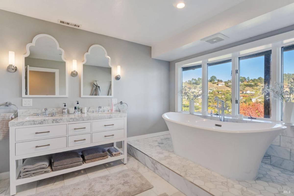 A bathroom with a double vanity, two mirrors, and a freestanding bathtub next to large windows.