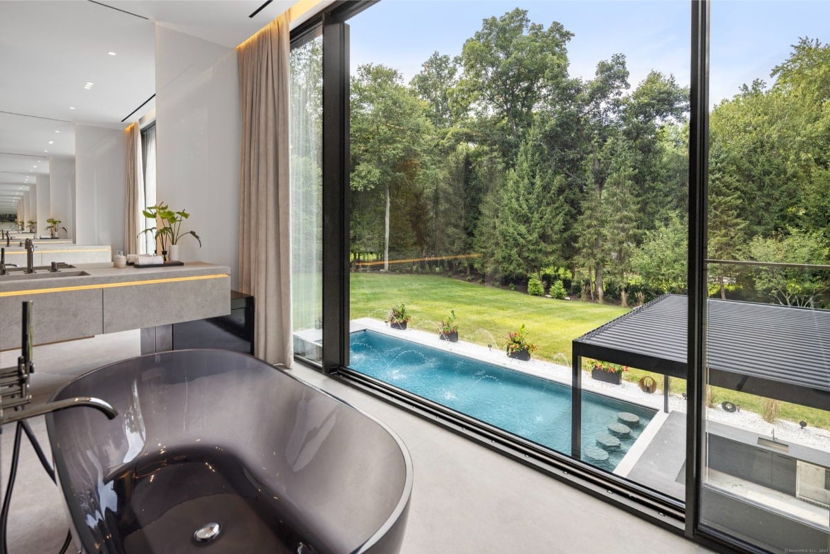 Sleek bathroom with dark bathtub positioned to maximize the view of an outdoor pool and lush green landscape through expansive windows.