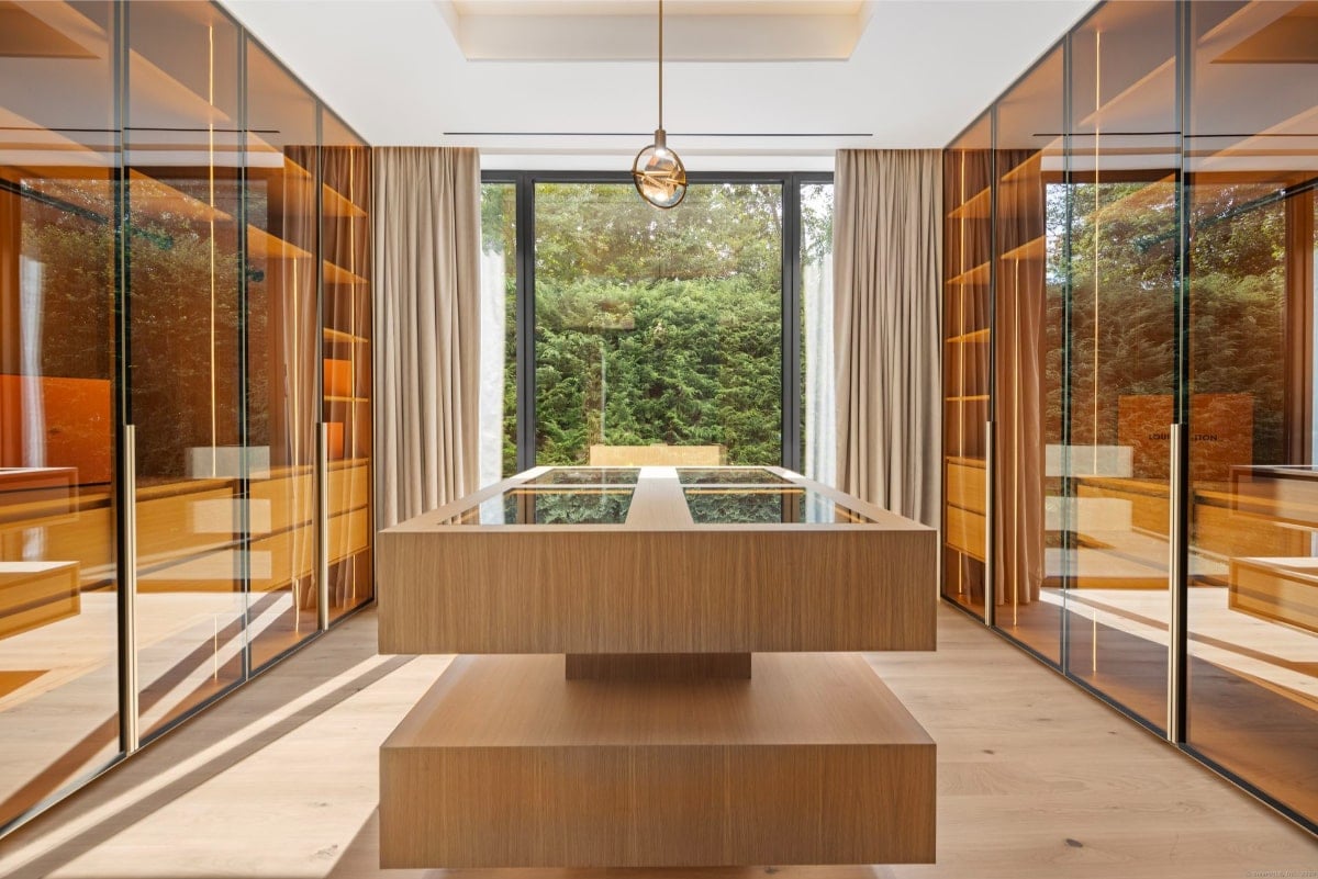 Walk-in closet with floor-to-ceiling, glass-fronted wardrobes. A large, low, rectangular piece of furniture with a glass top sits in the center of the room. Natural light streams in from large windows.