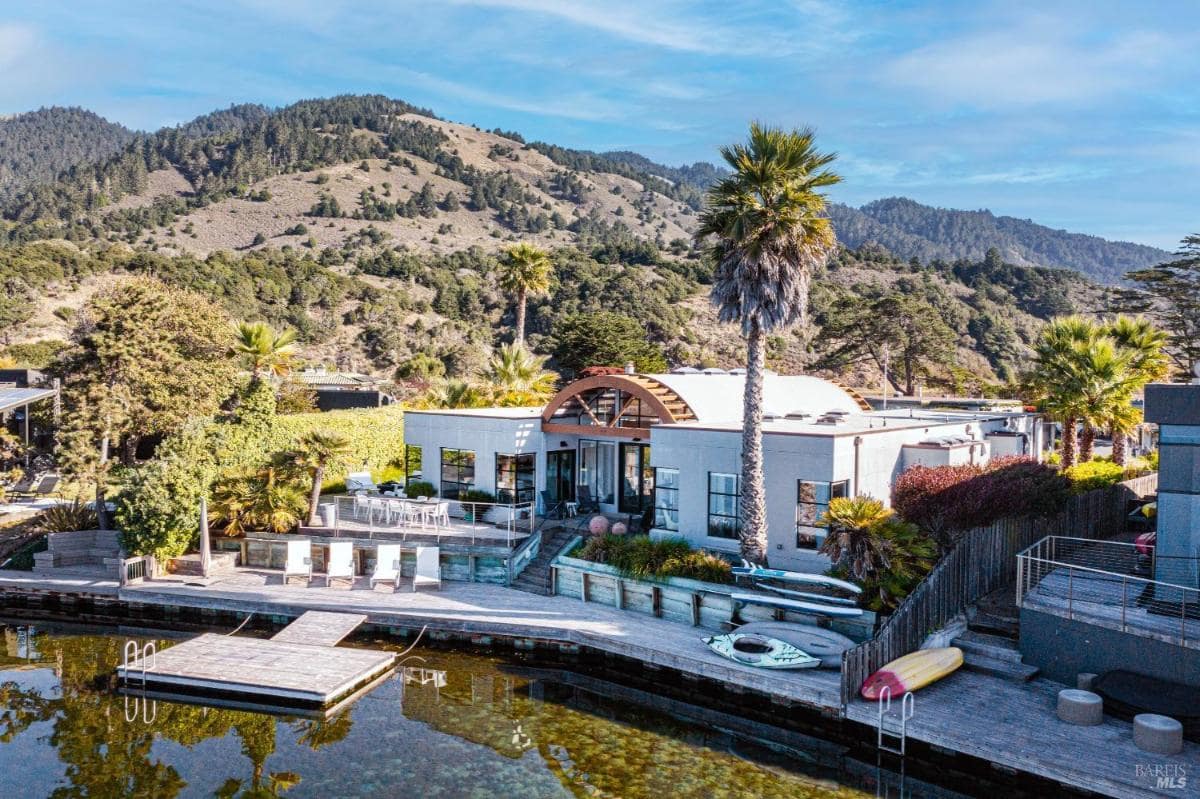 A contemporary house with a curved roof alongside a body of water, featuring outdoor seating and a mountain backdrop.