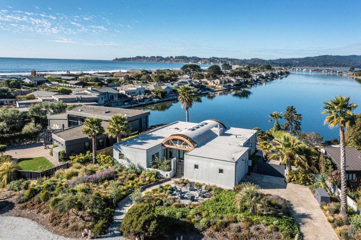 A modern house with a curved roof, overlooking a serene waterway and nearby ocean.