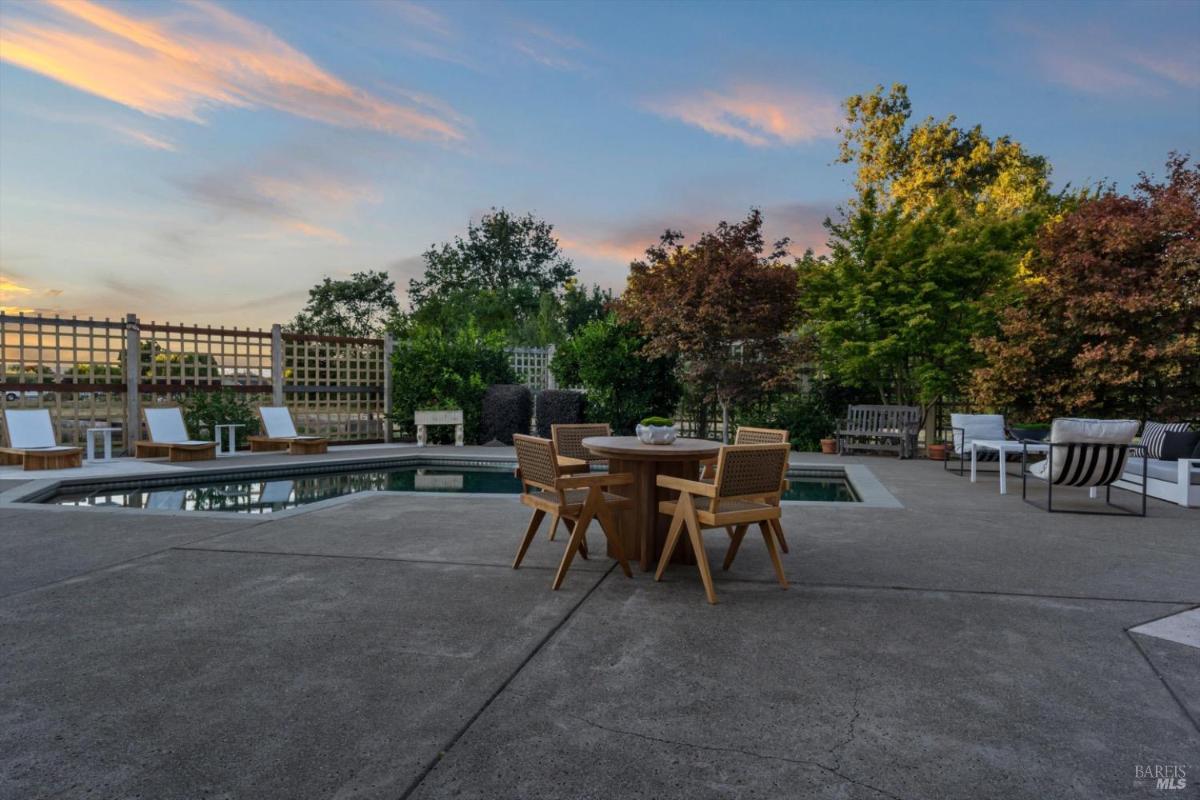 Patio with a round table, chairs, and a swimming pool surrounded by greenery.