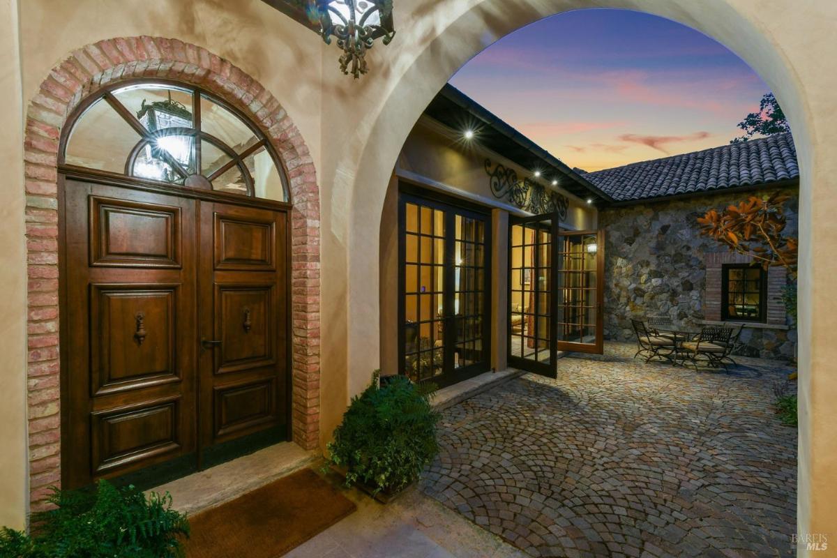 A wooden double-door entrance with brick arches, opening into a courtyard with cobblestone paving and patio furniture.