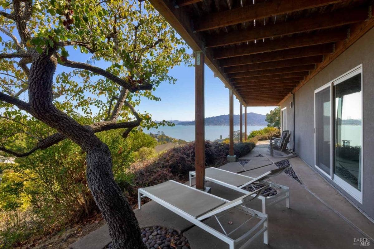 A patio area with lounge chairs, surrounded by greenery and overlooking a body of water and hills in the background.