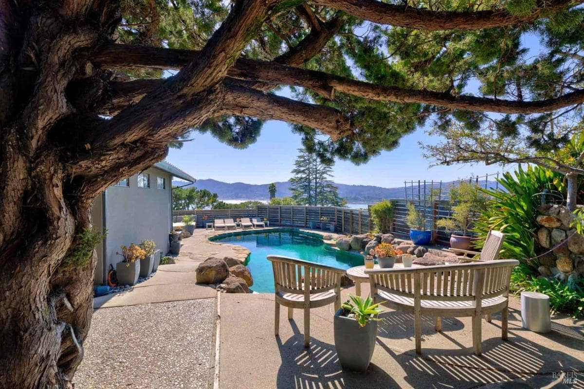 An outdoor space featuring a swimming pool surrounded by trees and plants, with wooden seating arranged for relaxation.