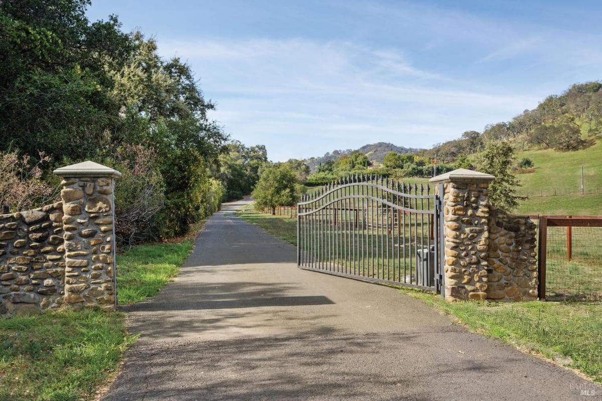 A gated driveway with stone pillars and an asphalt path, bordered by grass and trees.