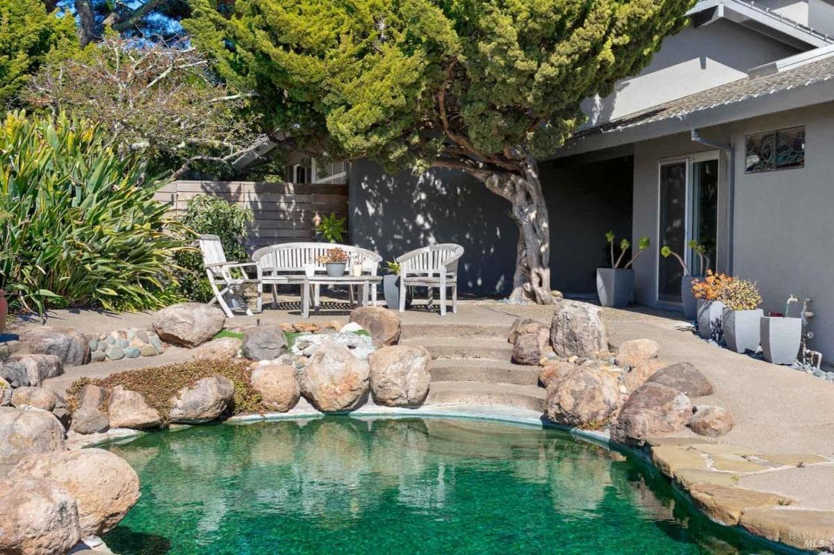 A small pool surrounded by rocks and a seating area near a house.