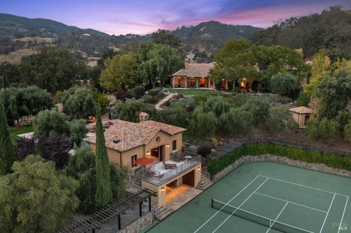 Overhead view of a multi-structure property with landscaped gardens, a tennis court, and surrounding hills.