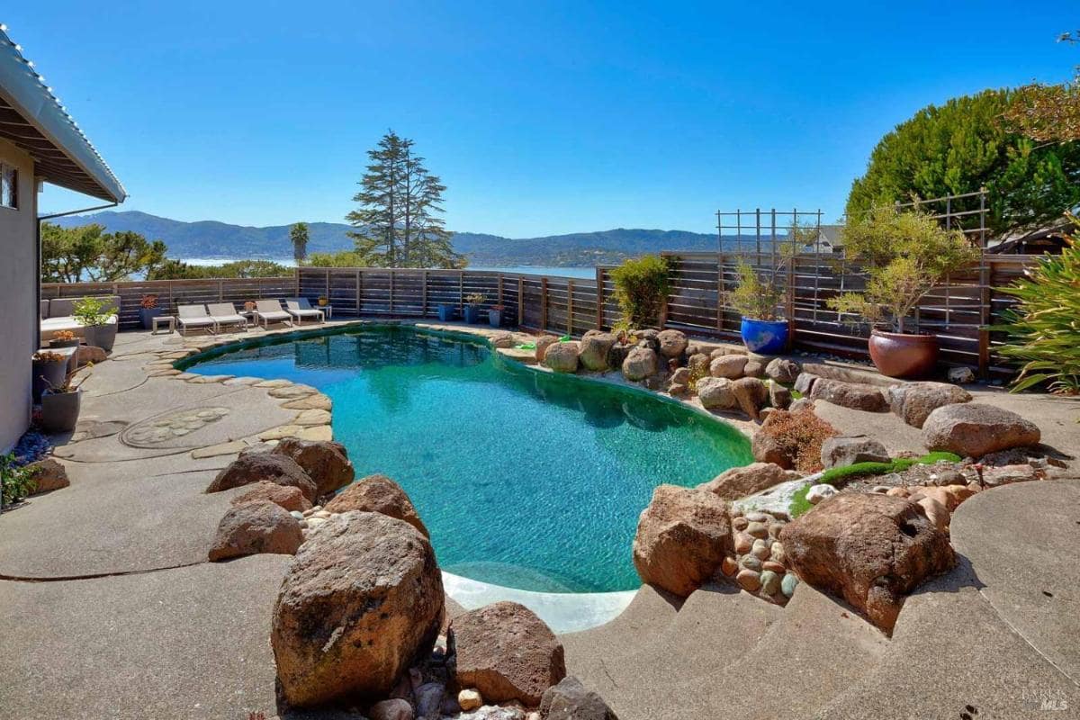 A pool area featuring natural stone accents, lounge chairs, and a scenic view of hills and trees under a clear blue sky.
