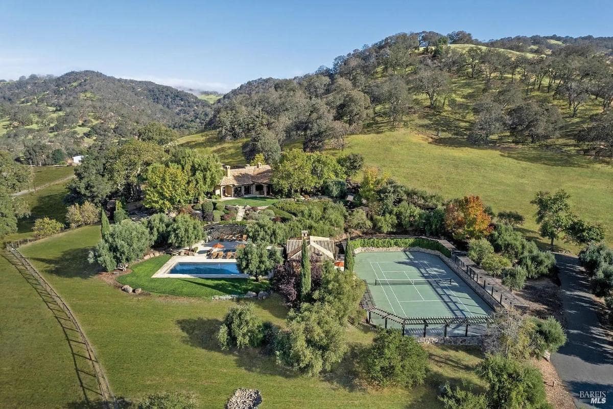Aerial view of a house with a swimming pool, tennis court, and gardens, set among rolling hills and open fields.