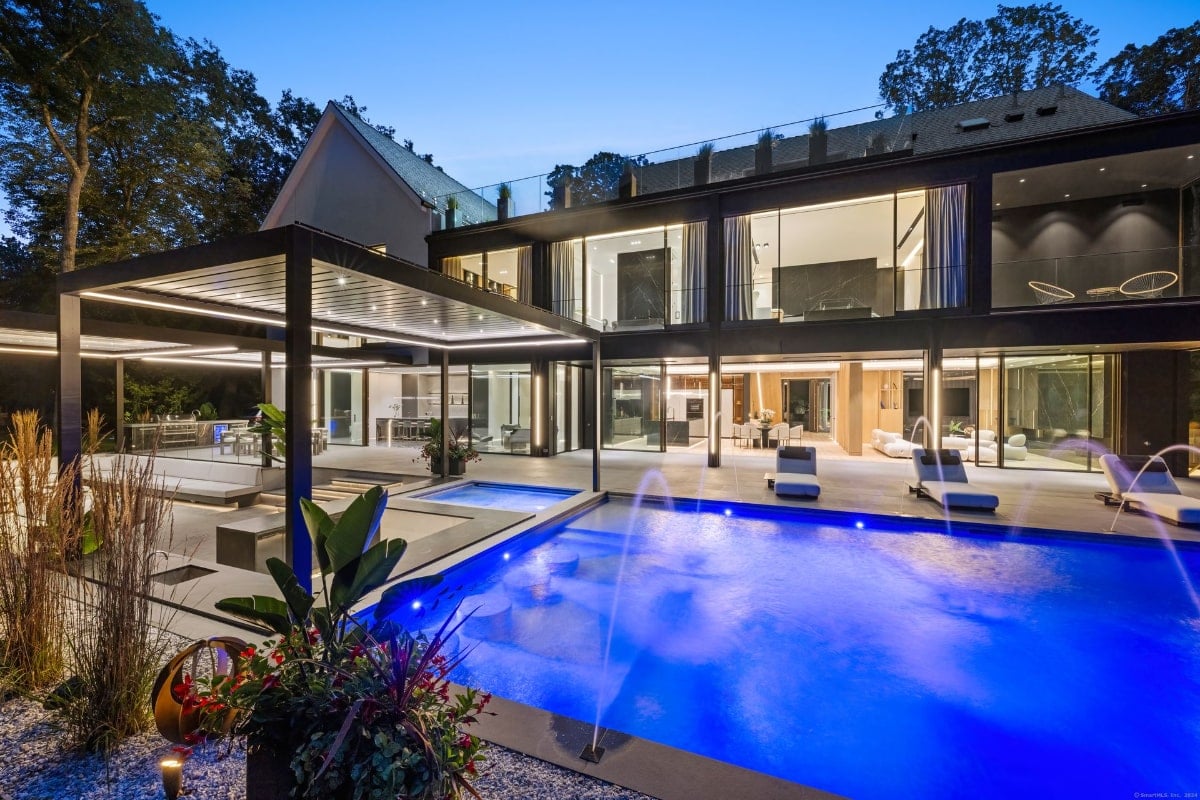 A luxurious mansion at night, with modern pool as the central feature. The mansion has a contemporary design with extensive glass windows, and there's a pergola-covered patio area adjacent to the pool. Water features add to the opulent feel.