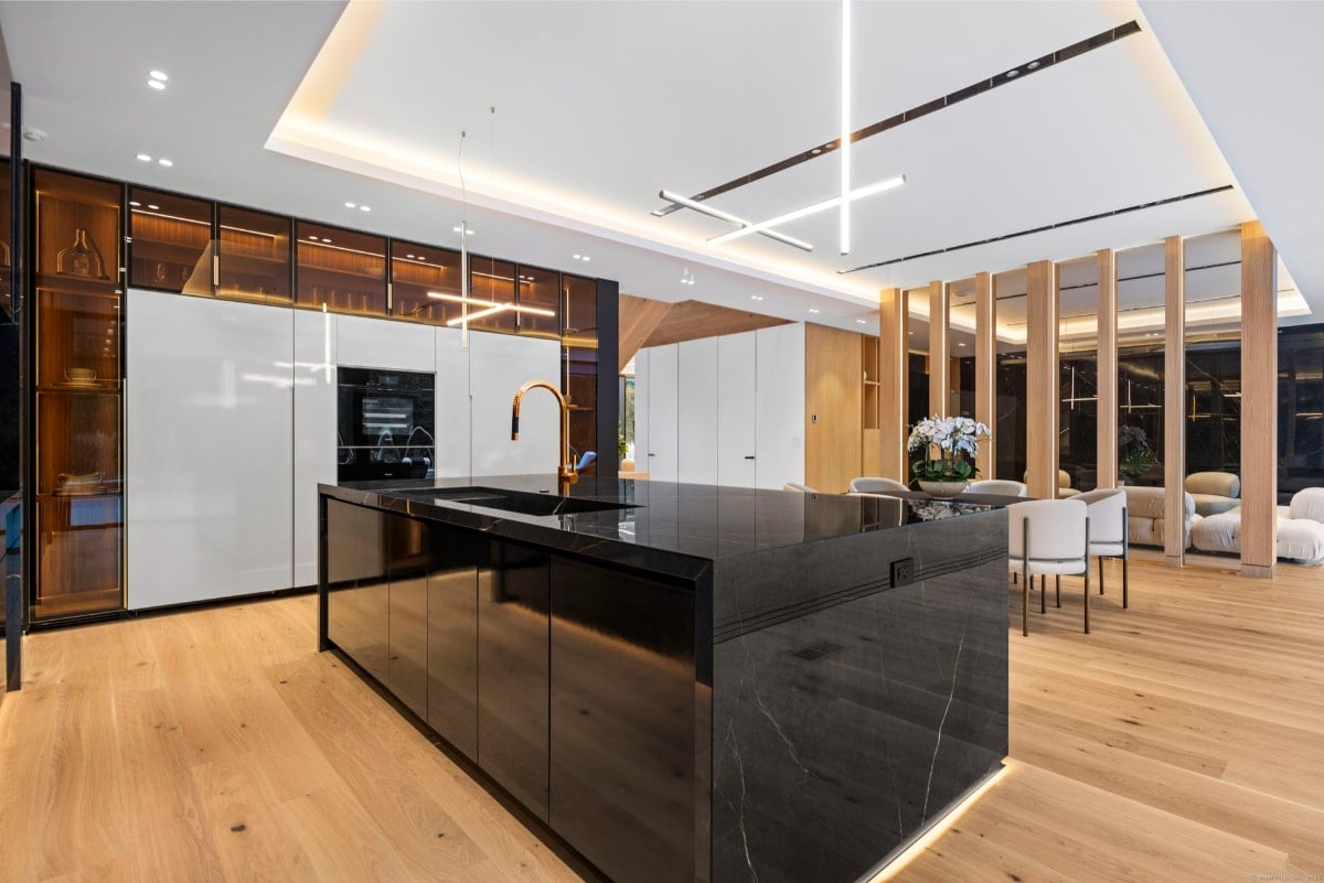Kitchen with a dark-colored island in the center. The island has a sleek, high-end appearance. Light-colored wood flooring and a dining area are visible in the background.