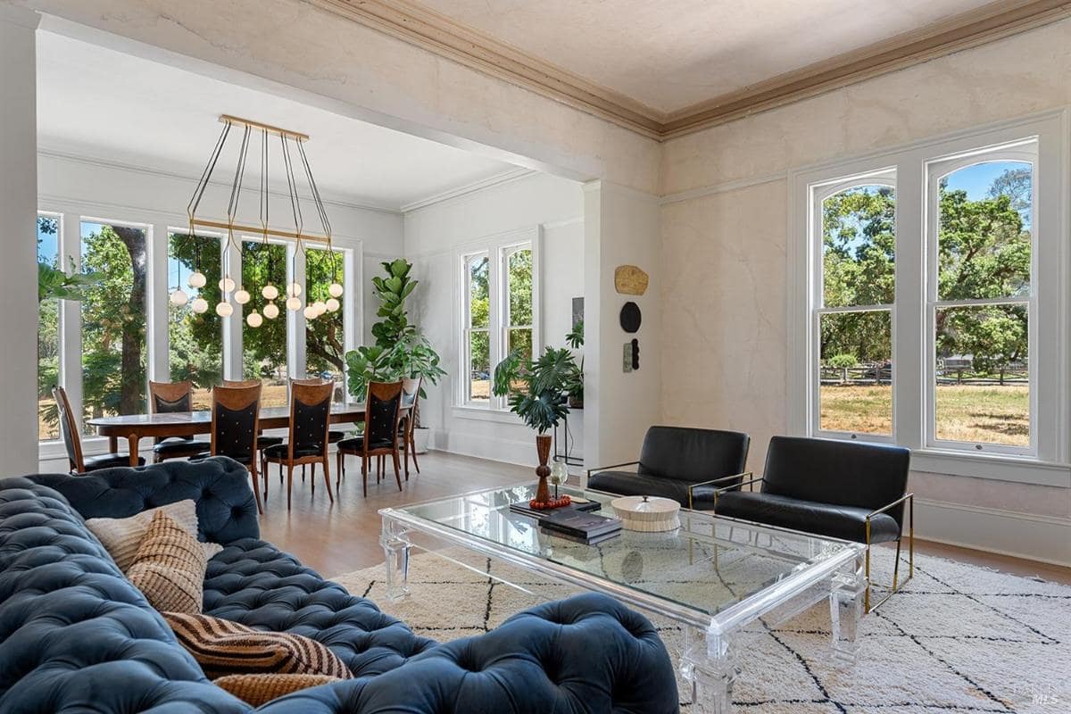 Living room with tufted blue sofa, glass coffee table, and large windows leading to dining area. 
