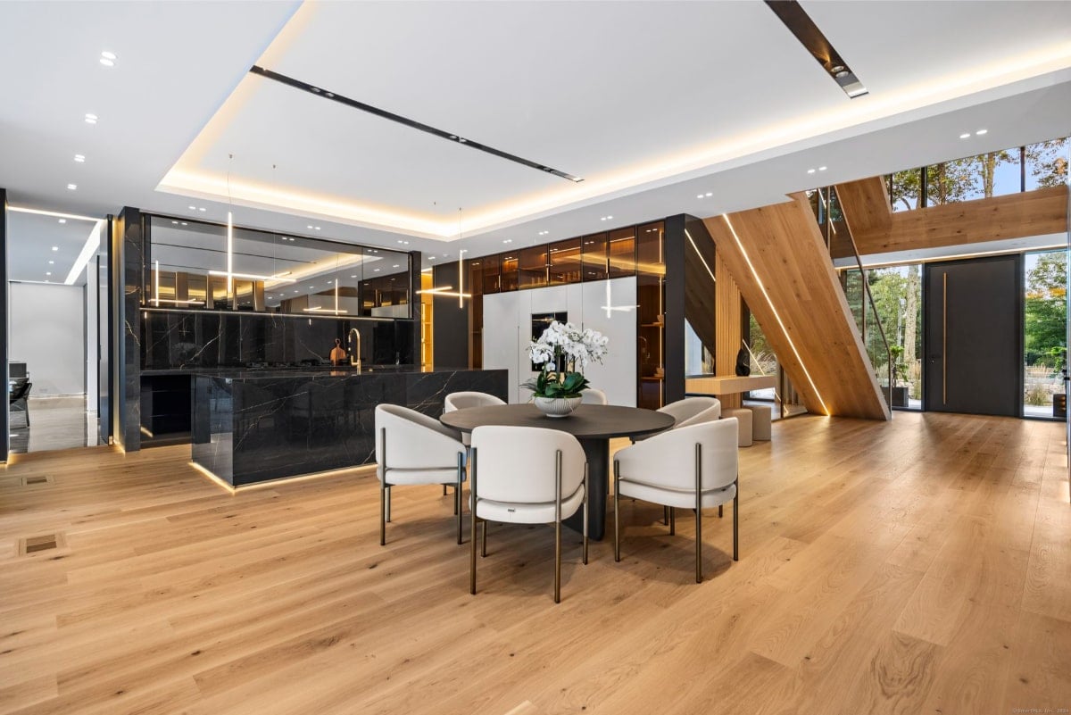 The dining area has a round table and four light-colored chairs. A sleek, dark kitchen island with marble countertops is visible in the background and flooring is light-colored wood.