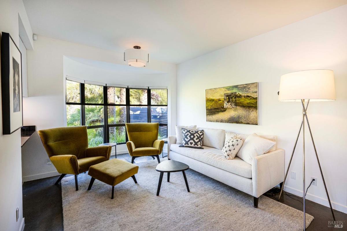 A small living area featuring a white sofa, two green armchairs, and a round coffee table near large windows. 