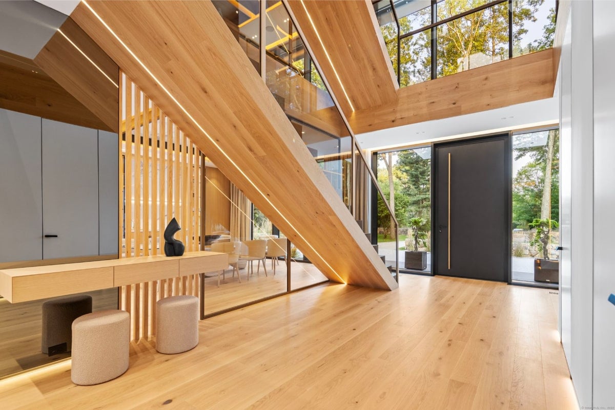 Light wood floors and a light-colored console table with stools are featured. A glass partition separates the foyer from a dining area beyond.