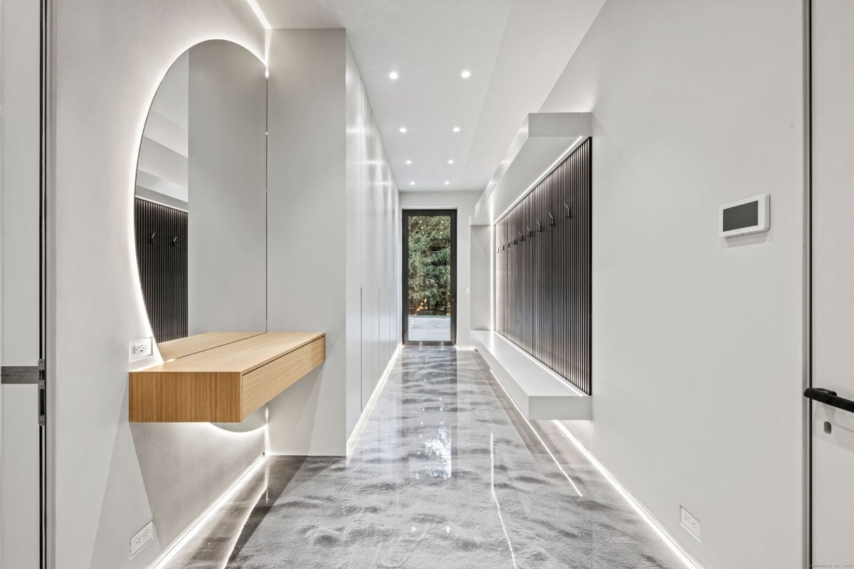 A narrow hallway with a modern design. There is a light wood console table under a large, curved mirror on one side, and a row of dark-colored storage units or lockers on the other.